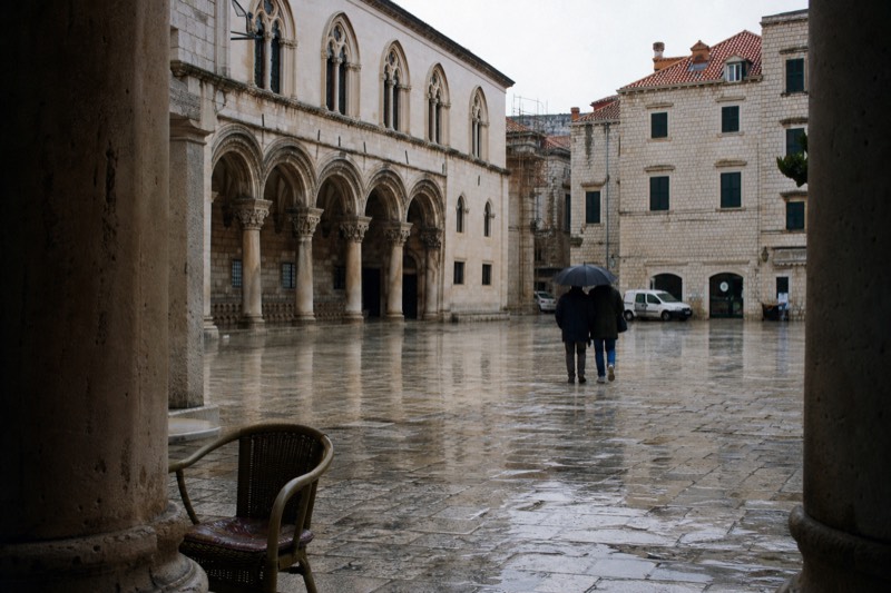 From inside a cool stone doorway Dubrovnik's Old Town square opens in soft grey winter rain, puddles doubling the arcade arches