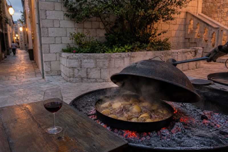 Evening in a Dubrovnik courtyard in Croatia, a domed iron peka lid lifts off an open fire, releasing steam over potatoes and meat in a black pan on glowing embers