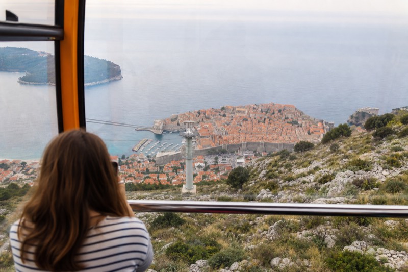 A yellow cable car cabin climbs above Dubrovnik, Croatia, on a spring afternoon, Mount Srđ scrub sliding past the glass, the walled old town and Adriatic spread below through soft haze