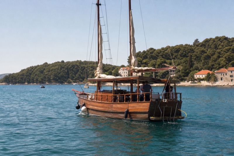 A wooden pirate-style karaka with furled sails glides across turquoise water between pine-covered islets of the Elaphiti Islands, Croatia, midday glare