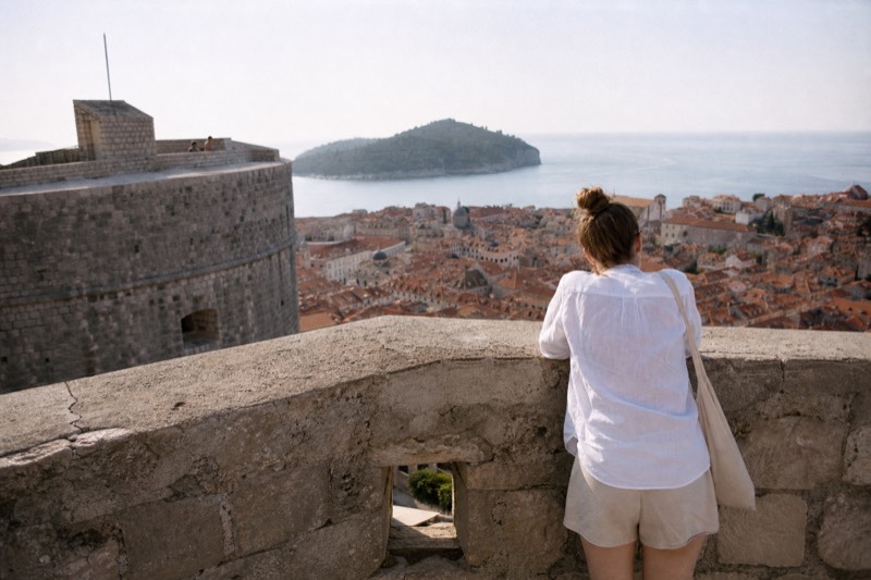 Early summer morning on the round stone ramparts of Minčeta Fortress in Dubrovnik, Croatia, terracotta rooftops dropping away below; a visitor from behind leans on warm limestone parapet