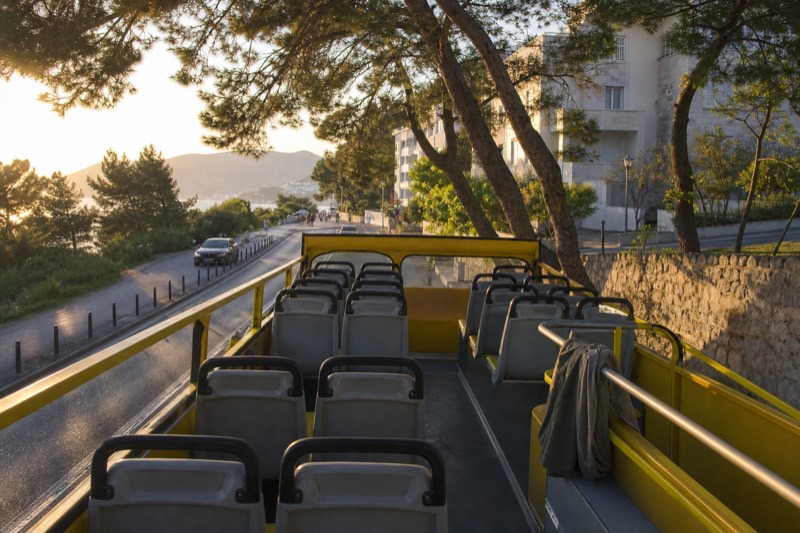 Late afternoon in spring at a Babin Kuk stop in Dubrovnik, Croatia, the open upper deck of a yellow sightseeing bus seen from behind and above, empty seats catching low sun