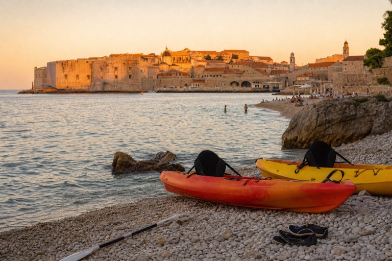 At golden hour kayaks rest on Banje beach's pebbles, Dubrovnik's city walls glowing orange across the water