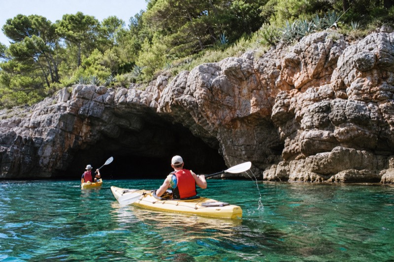 Sea kayakers paddling past the dark rocky entrance of Betina Cave beneath Lokrum Island near Dubrovnik, Croatia, morning sun on clear turquoise water, a paddle blade dripping