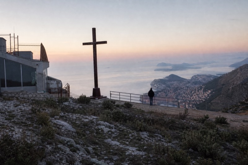 The Mount Srđ summit station above Dubrovnik, Croatia, first light, dry limestone scrub and a rusted cross silhouette