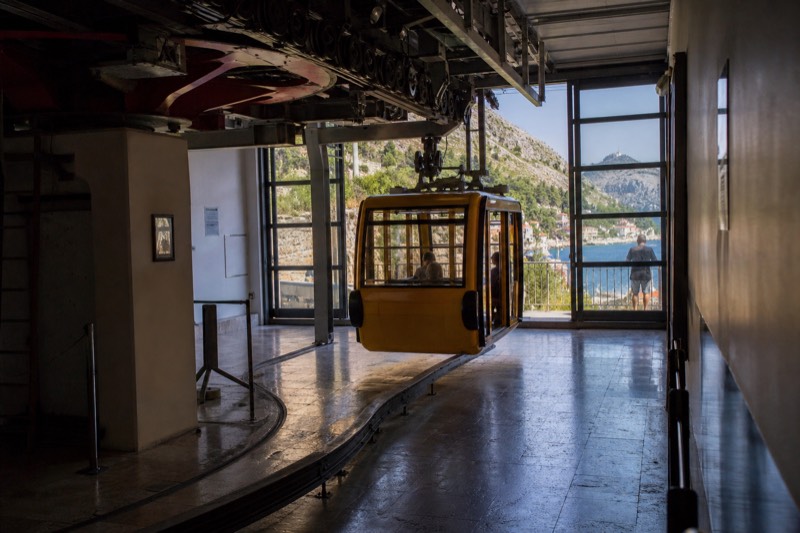 Inside the lower cable car station in Dubrovnik, Croatia, a yellow cabin gliding toward the glass doors, steel cables and pulleys overhead, morning light catching scuffed tiled flooring