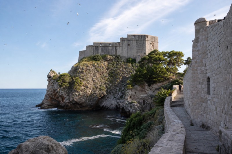 Late morning sun raking across Fort Lovrijenac's weathered limestone blocks on its sea rock below Dubrovnik's walls, a distant visitor paused on the steep stair, swifts overhead