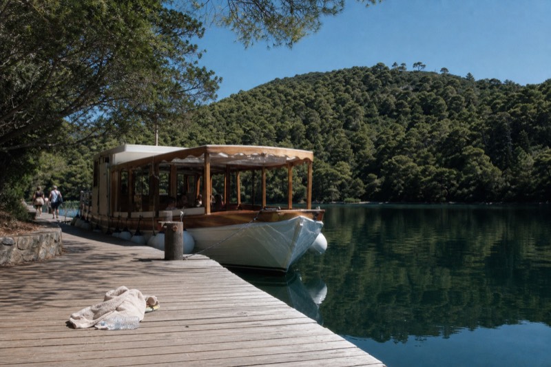 A tour boat moored at a wooden jetty on Mljet's saltwater lake in Croatia, dense green forest reflected in still water, summer noon, a folded towel and empty water bottle left on the planks