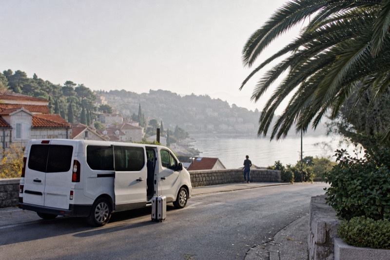 A white shared transfer van waits on a steep coastal road above Cavtat, Croatia, early morning, silver suitcase by the open sliding door, sea haze over the pine-topped hillside