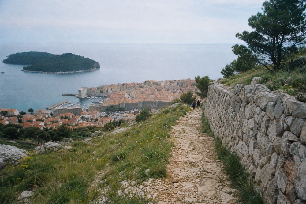 A dry stone trail winding down the sunlit slope of Mount Srd with the walled city below