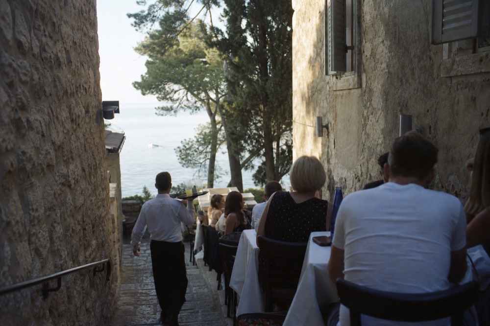 Waiter carrying a tray of drinks between closely set tables at an evening restaurant terrace in a narrow stone alley