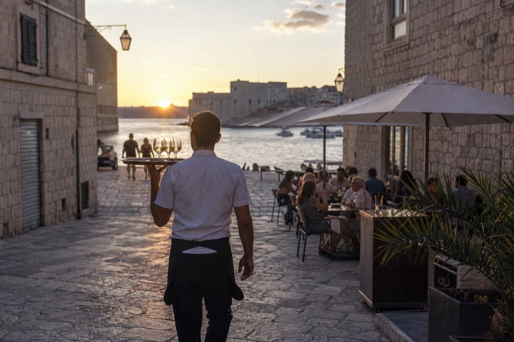 A waiter carrying a tray across a narrow stone street with outdoor tables