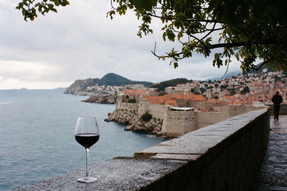 A wine glass of red wine on a stone ledge with the old city walls and sea behind it