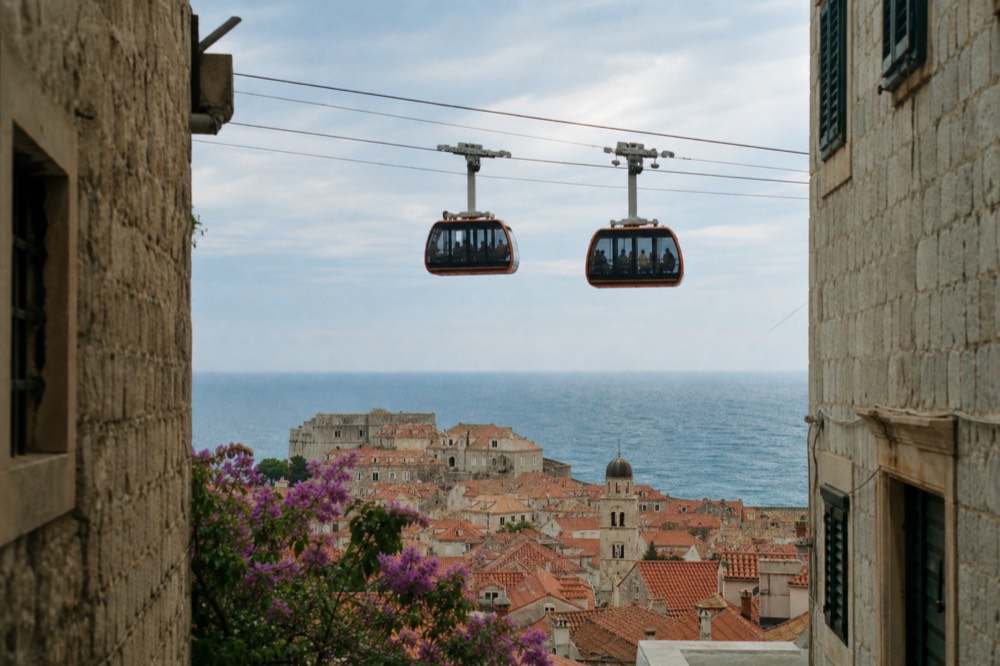 Two cable car cabins passing each other above the tiled roofs of Dubrovnik with the Adriatic sea behind