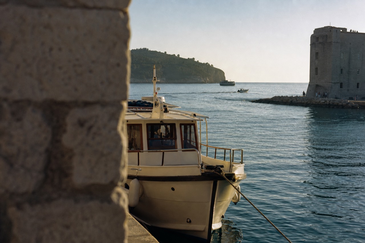 A small passenger boat moored in the Old City Harbour of Dubrovnik with Lokrum island visible across the water