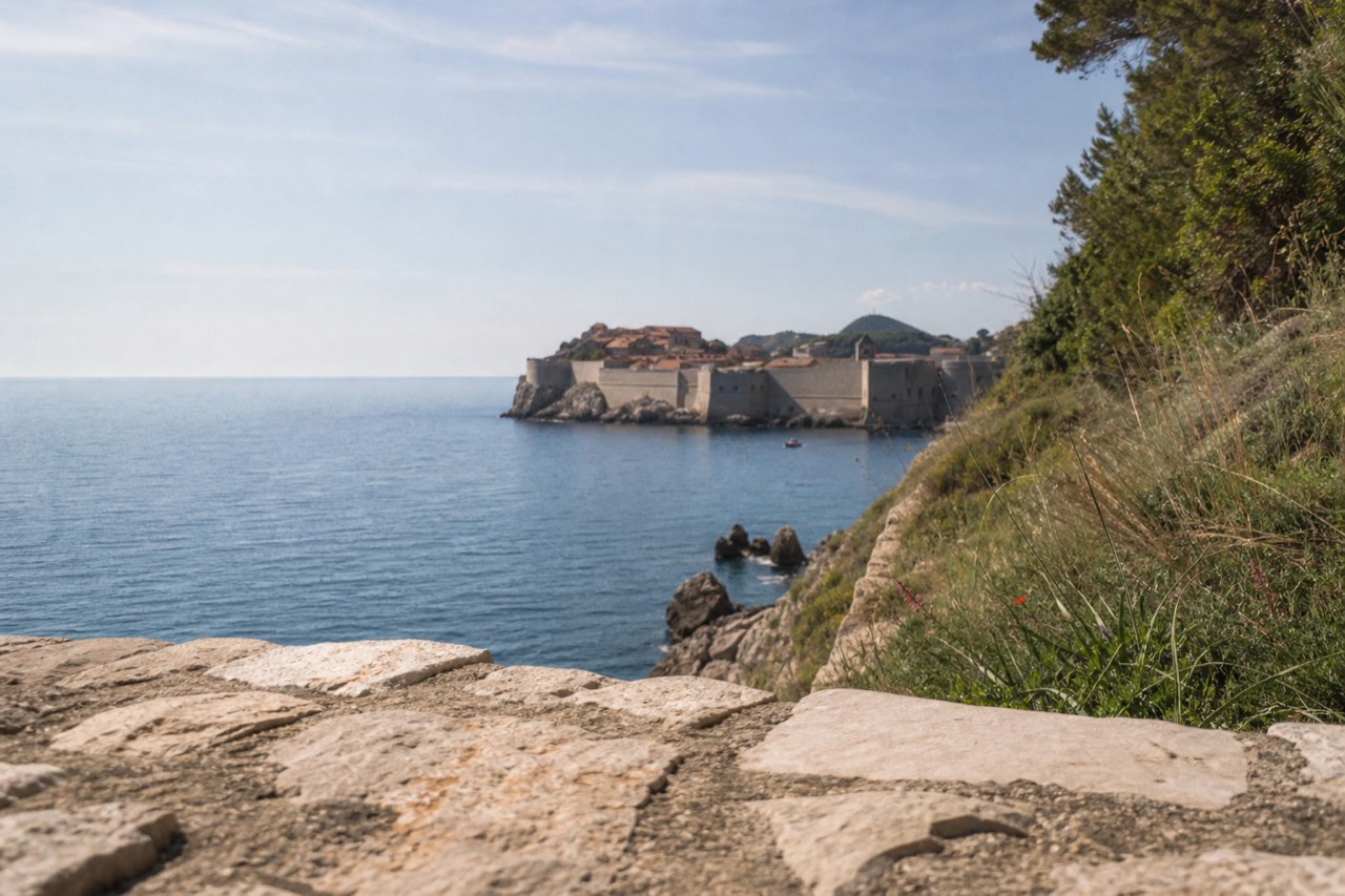 Sunlit Adriatic coastline below Dubrovnik with calm blue water and the walled Old Town in the distance