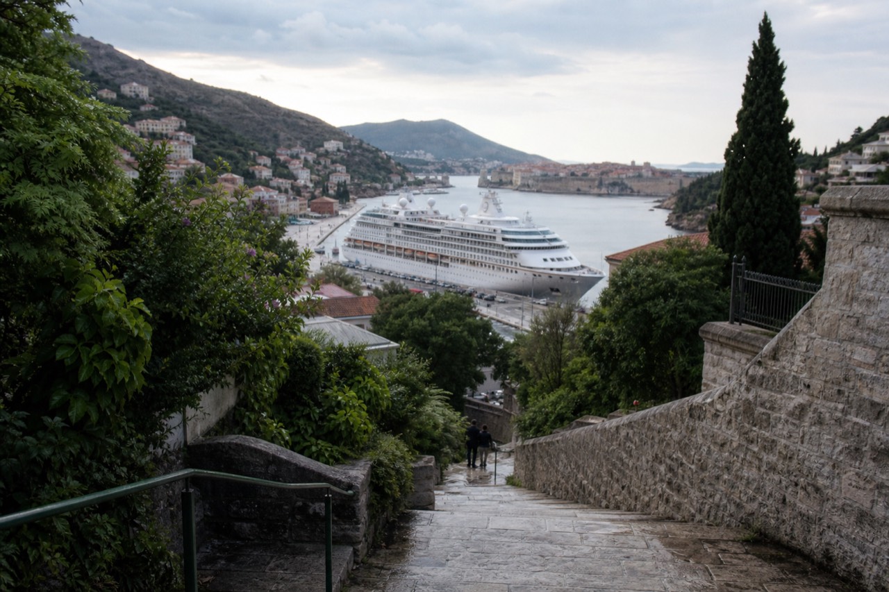A cruise ship docked at a port with a walled old town and terracotta roofs in the distance