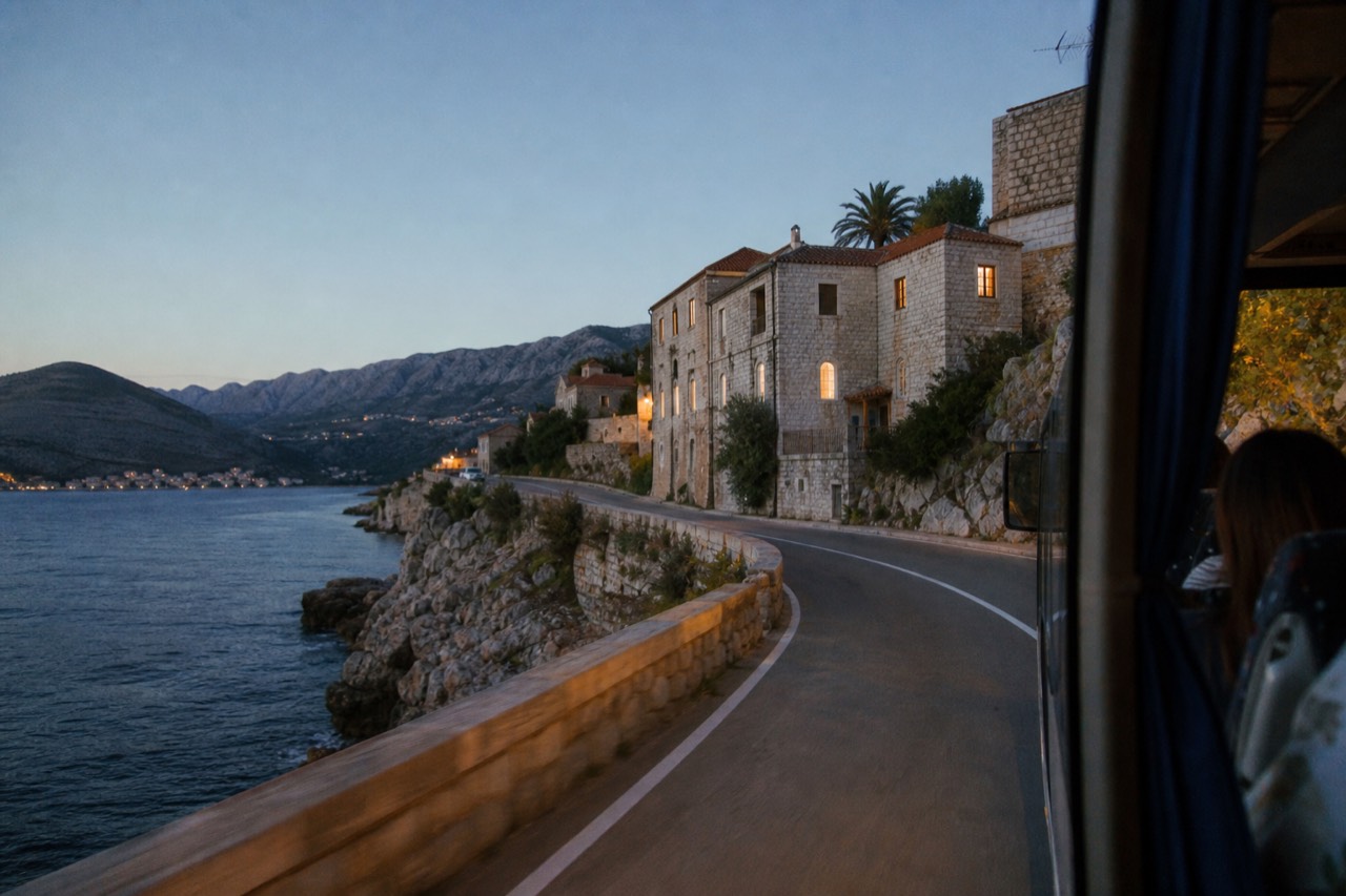 A scenic coastal road winding along the Adriatic Sea with mountains in the background, taken from a bus window.