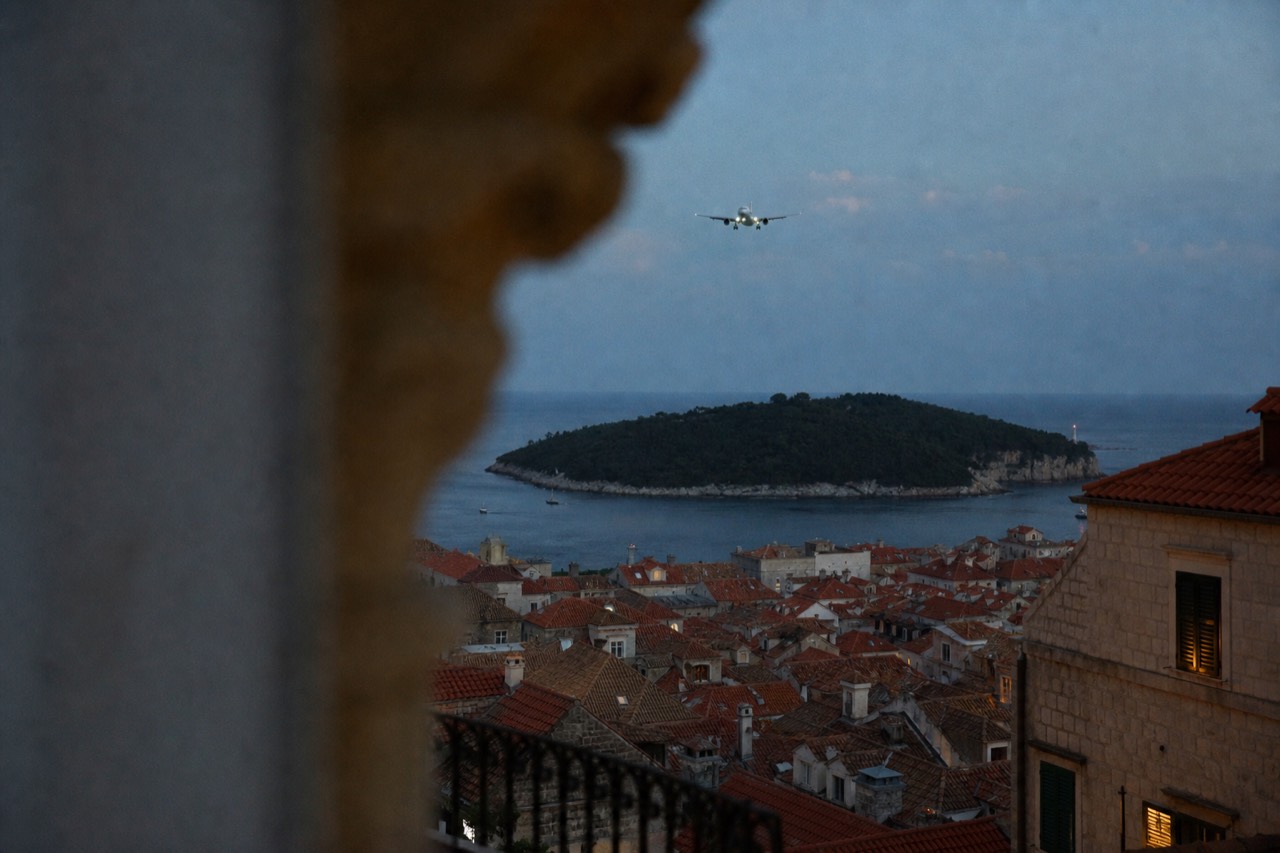 A plane descending over a walled Adriatic coastal town with terracotta rooftops and a green island offshore