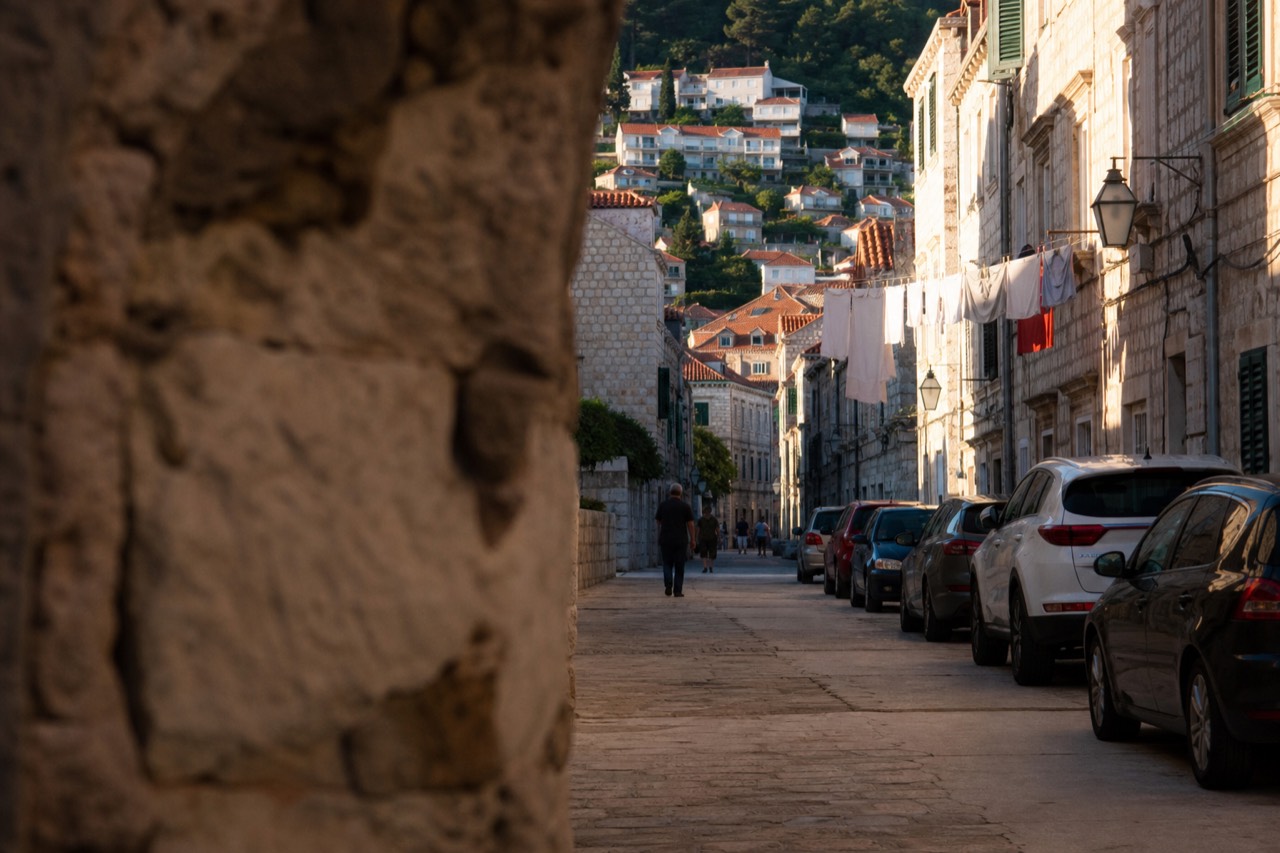 A narrow stone street in Dubrovnik with parked cars lining one side and laundry hanging above