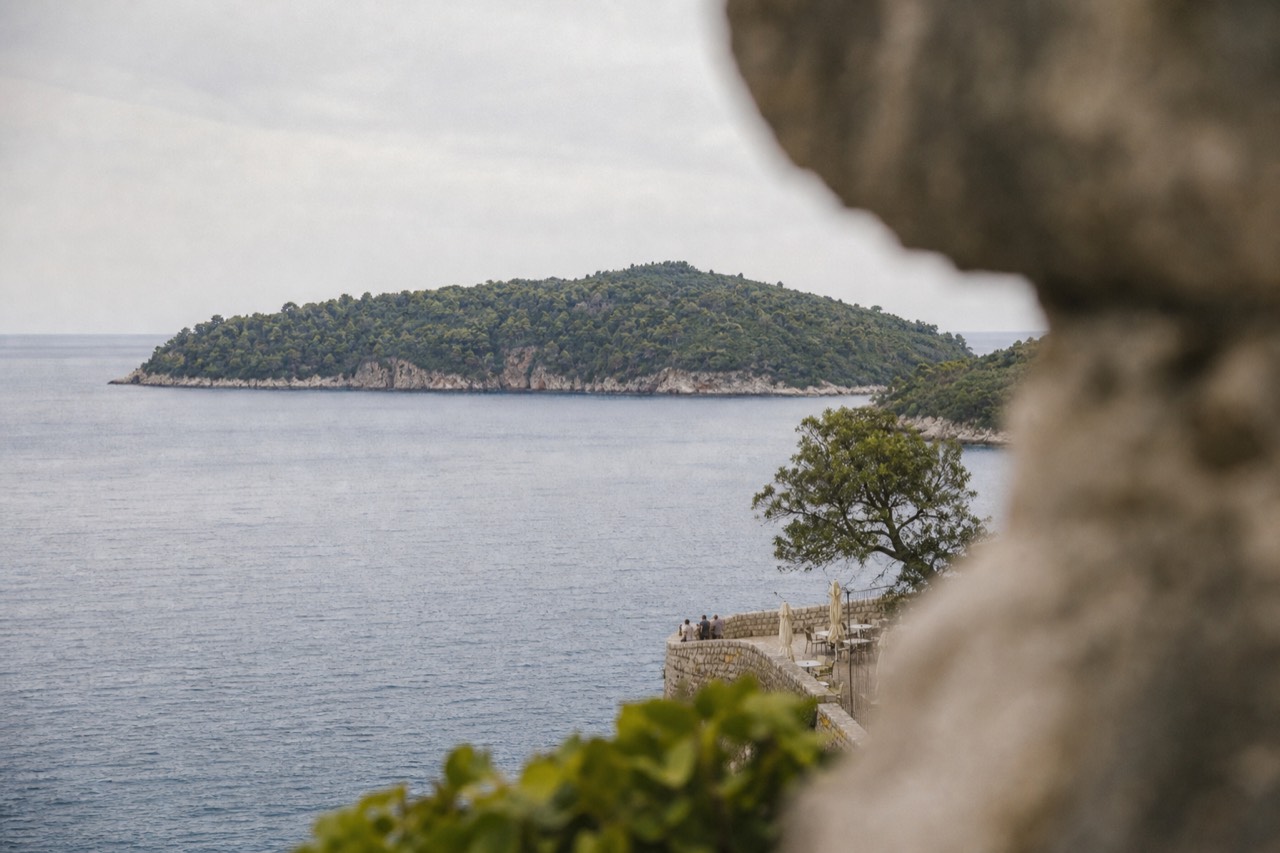 A wooded green island rising from calm blue Adriatic water a short distance from a walled seaside town.