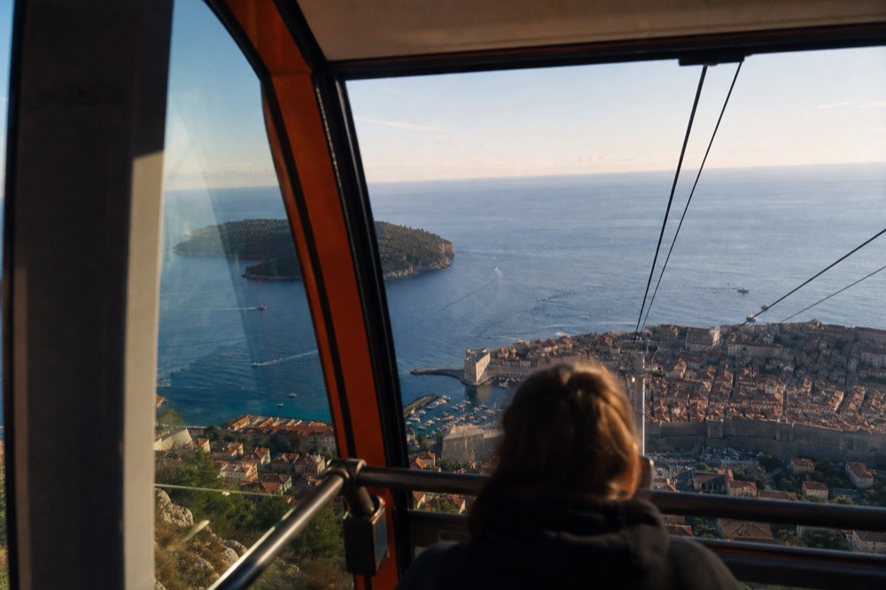 Cable car cabin ascending above Dubrovnik's Old Town with the Adriatic Sea and Lokrum Island in the background.