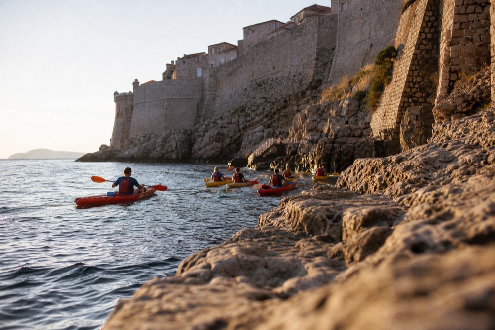 A group of sea kayaks paddling along the stone cliffs below Dubrovnik's city walls