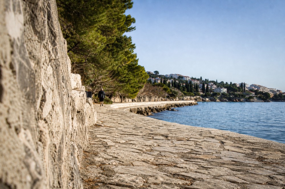 A quiet stone promenade curving around the Lapad bay with pine trees and calm water