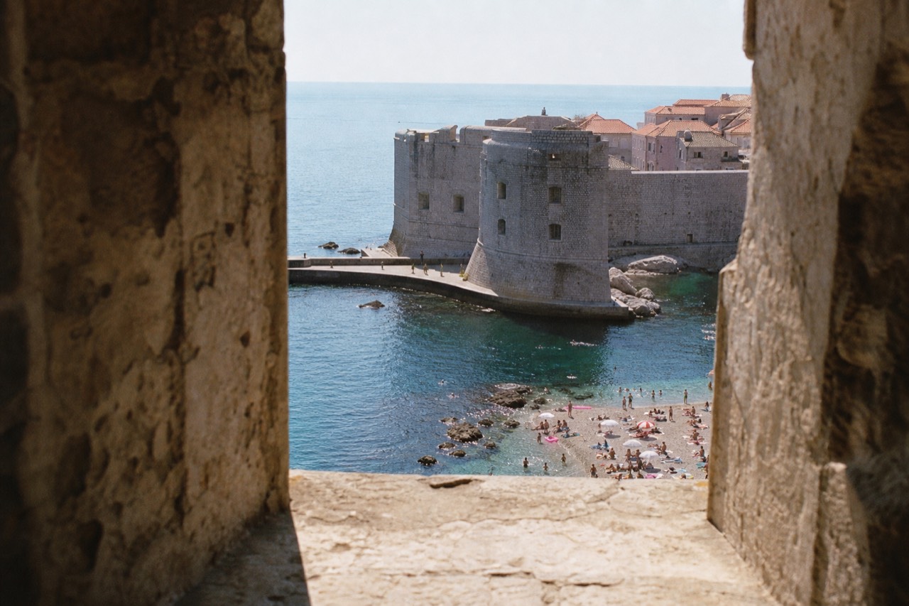 A stone walled old town beside calm blue Adriatic water with a small swimming beach in the foreground