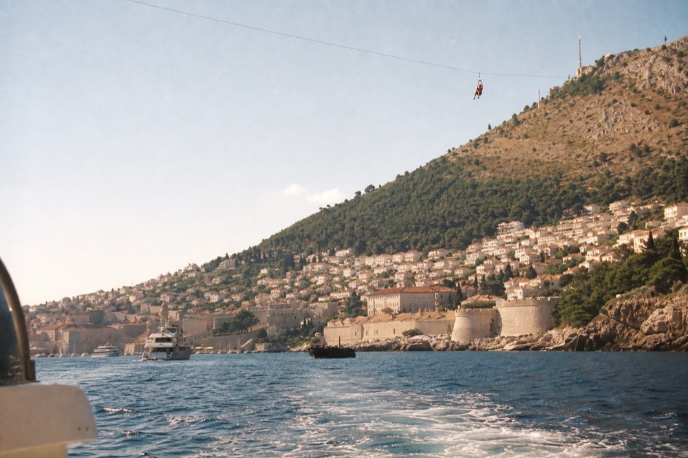 A person in a harness riding a zipline cable above steep hillsides with the sea in the distance