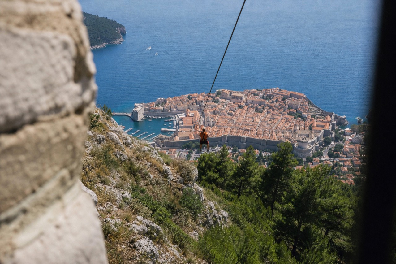 A zipline cable stretching over pine-covered slopes above the walled city and the blue Adriatic sea