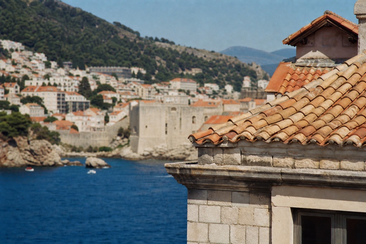 A stone seaside town with terracotta roofs and calm blue water below wooded hills