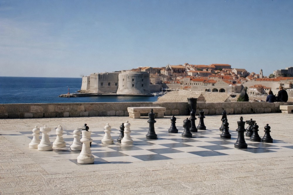 A giant stone chessboard with large pieces on a paved square beside old city walls