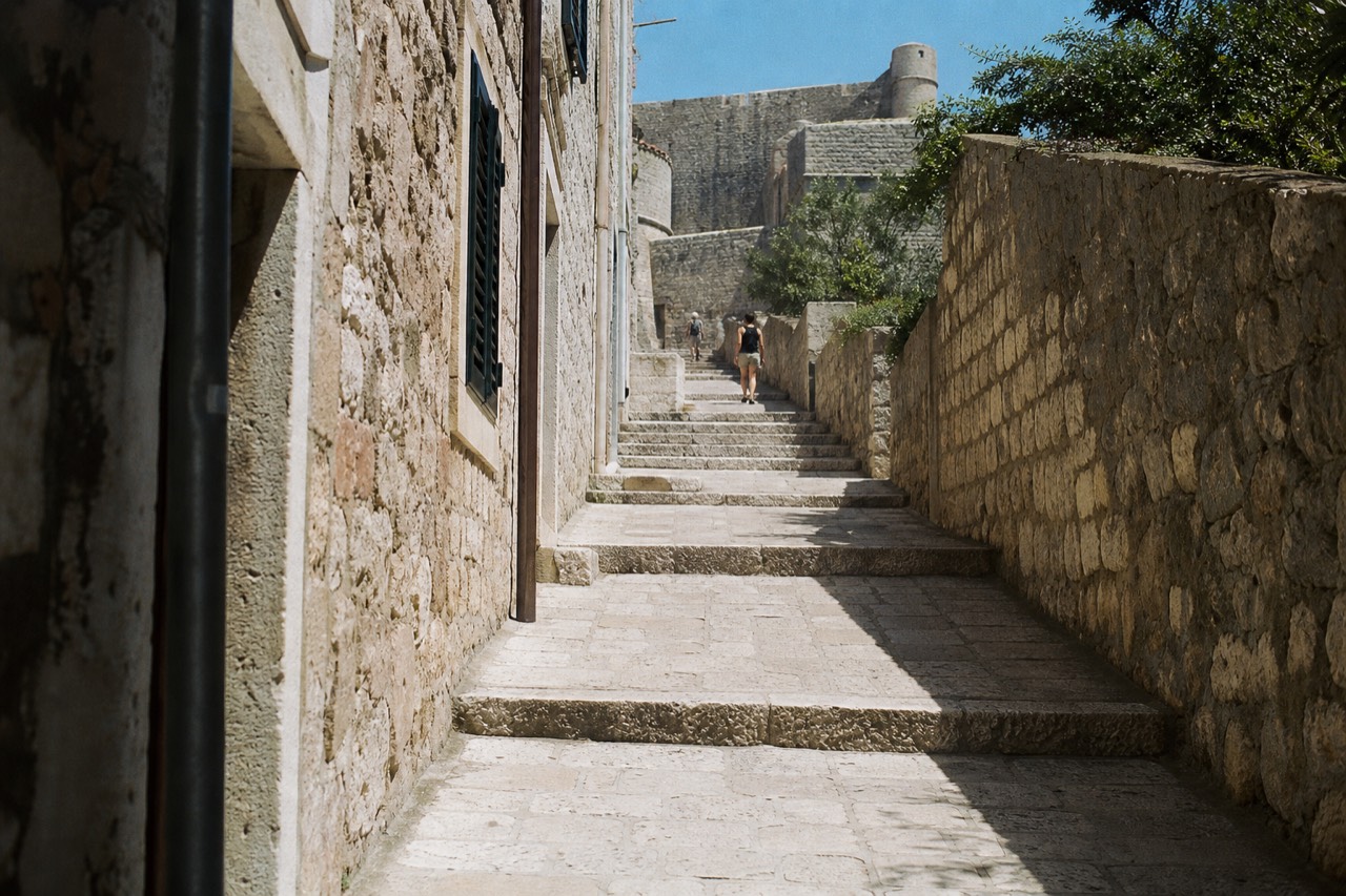 Sunlit limestone street inside the walled Old Town of Dubrovnik with stone staircases rising toward the city walls