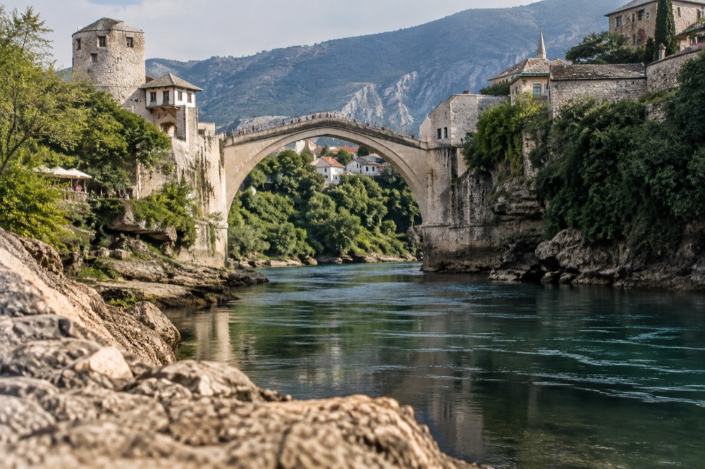 A stone bridge arching over a river gorge with a green valley and old buildings on the cliffs