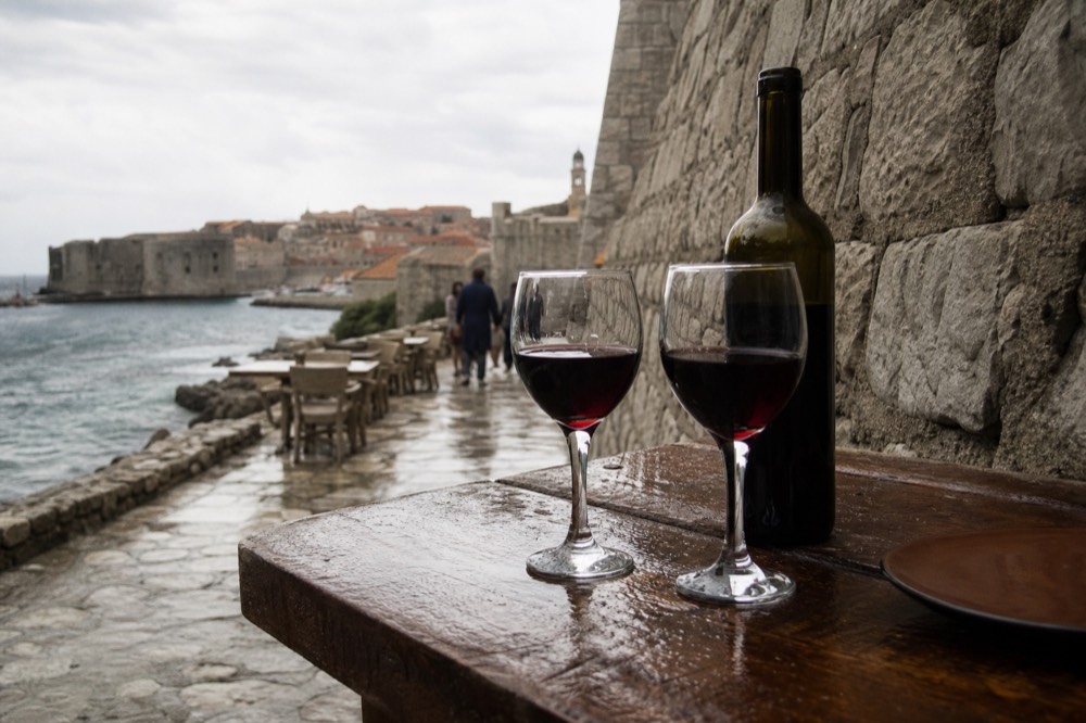 A bottle of red wine and two filled glasses on a rustic wooden table beside a stone wall