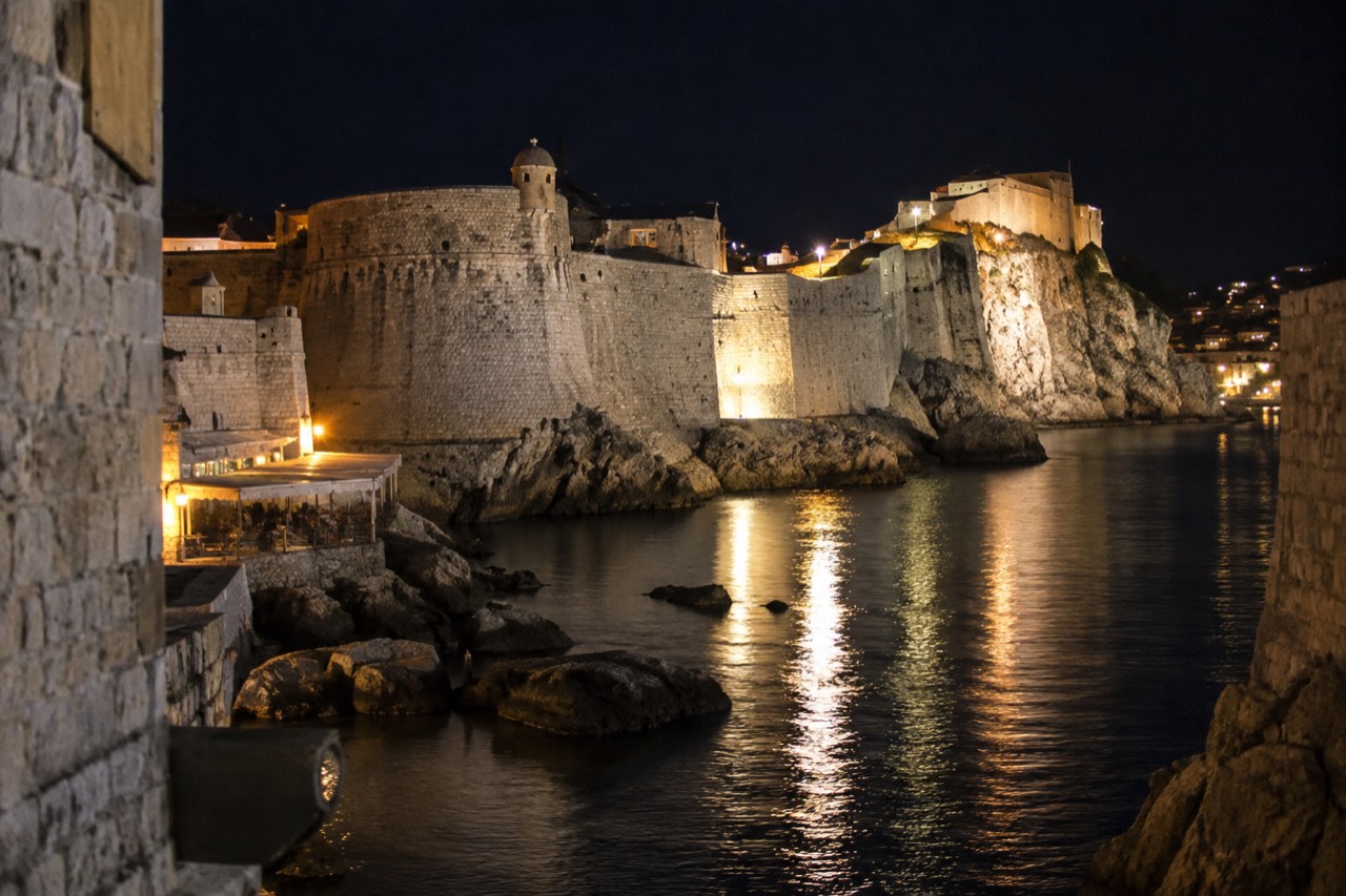 Stone fortress walls above a dark harbour with lights reflecting on the water at night
