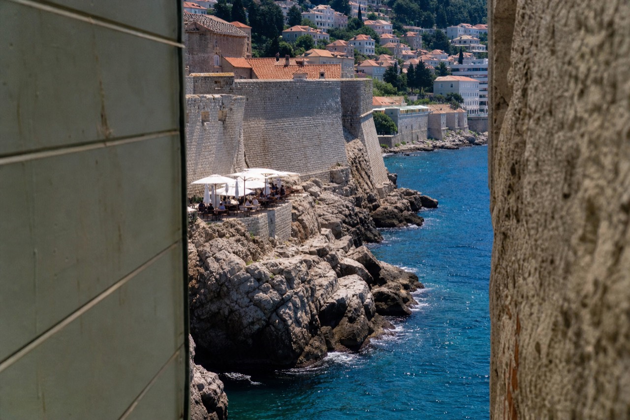 A small terrace with tables and umbrellas on rocks above the sea below stone walls