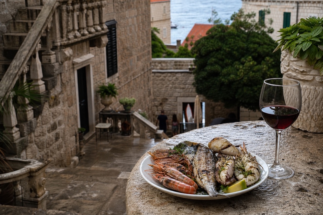 A plate of grilled fish and shellfish on a stone table beside a glass of red wine in an old stone courtyard