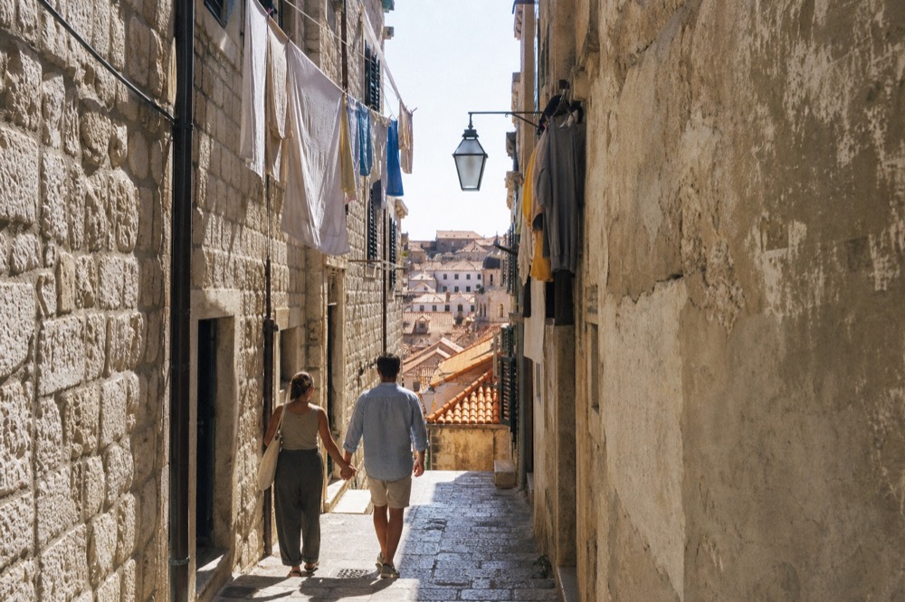 A couple walking down a narrow stone alley in Dubrovnik Old Town, surrounded by limestone buildings and hanging laundry
