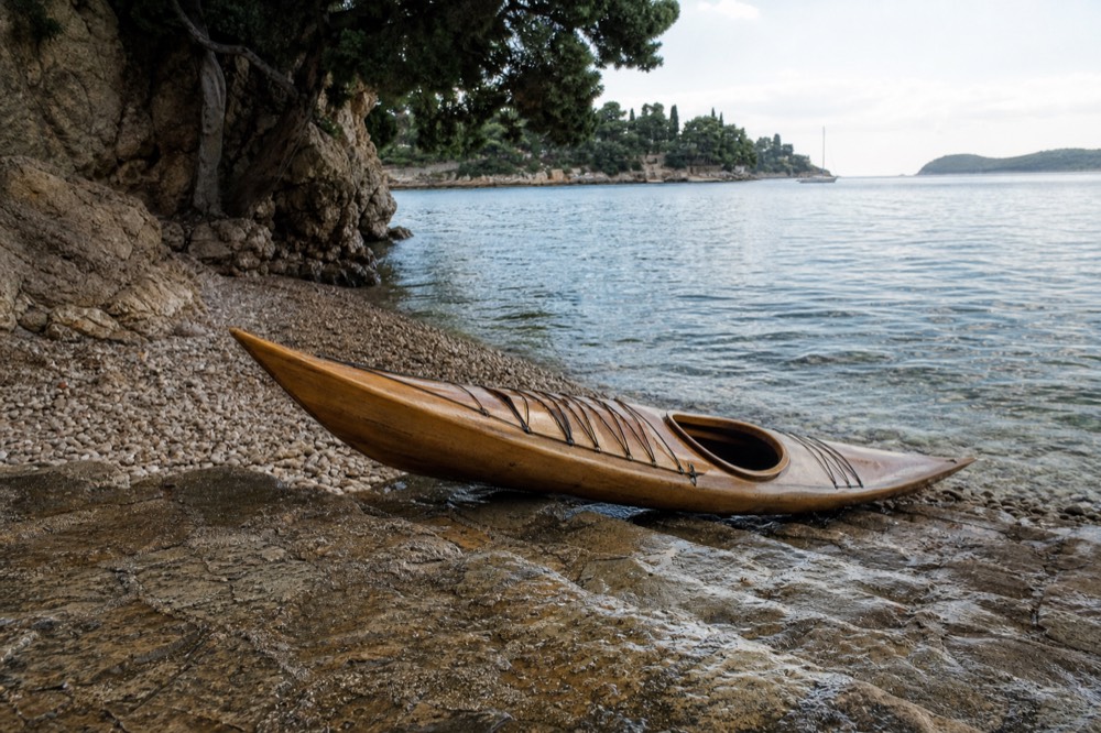 A wooden kayak resting on smooth pebbles beside clear shallow water