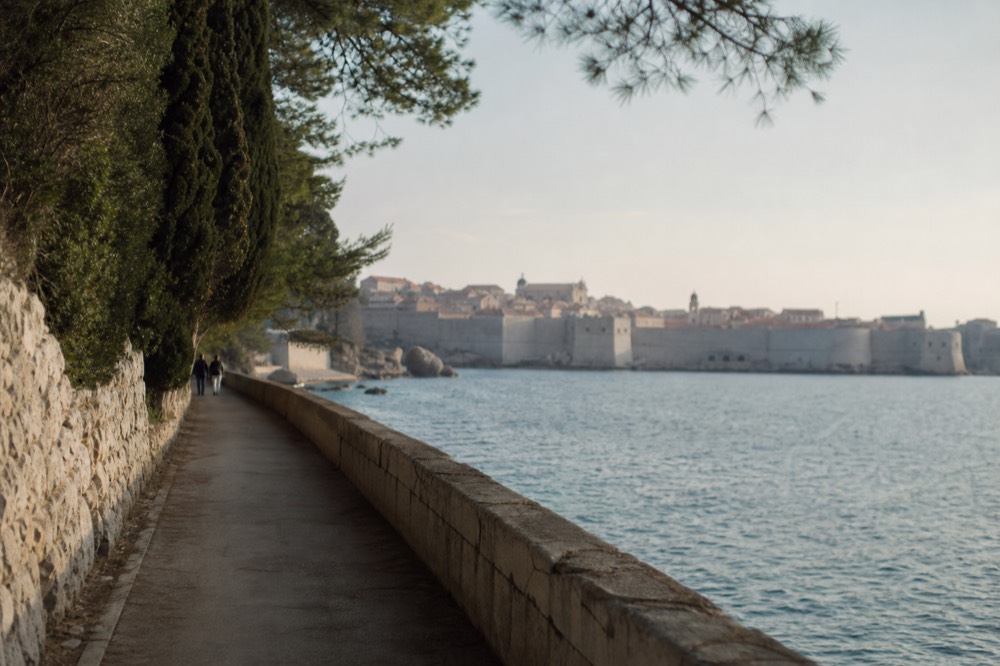A waterfront promenade beside the sea leading toward the walled city in the distance