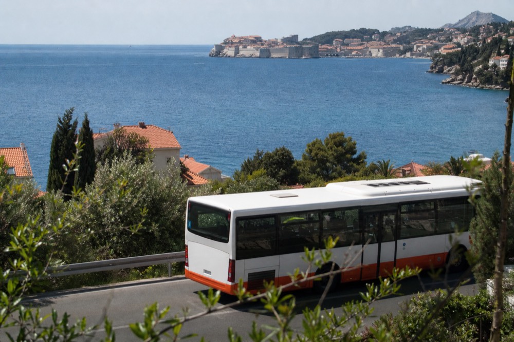 A city bus on a coastal road with the sea and a walled town in the distance