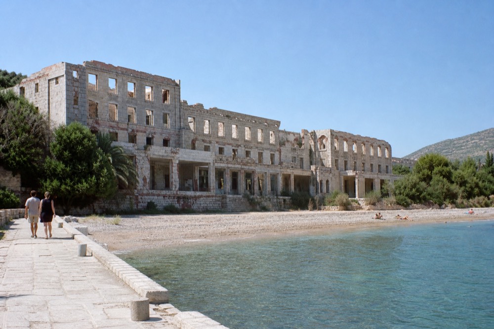 Ruin of a multi-storey hotel with empty window frames above a calm pebble beach