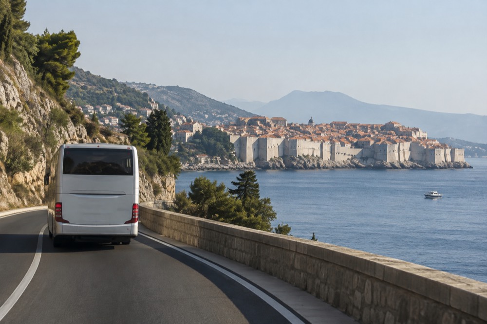 A bus driving along a coastal road with the walled city visible across the water