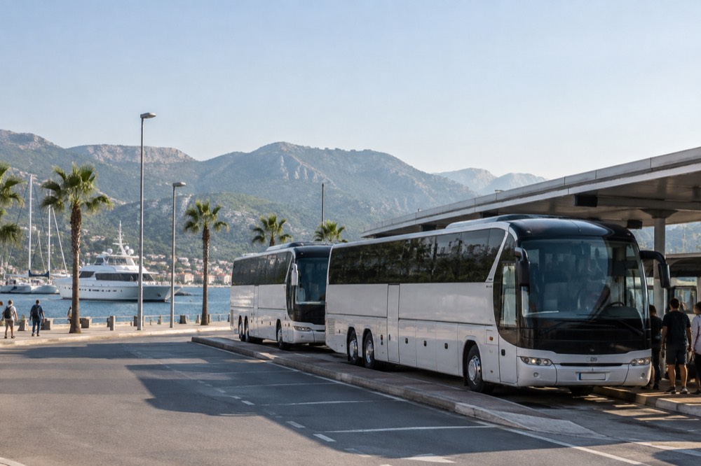 Buses parked at a station platform beside a harbour with boats moored in the background