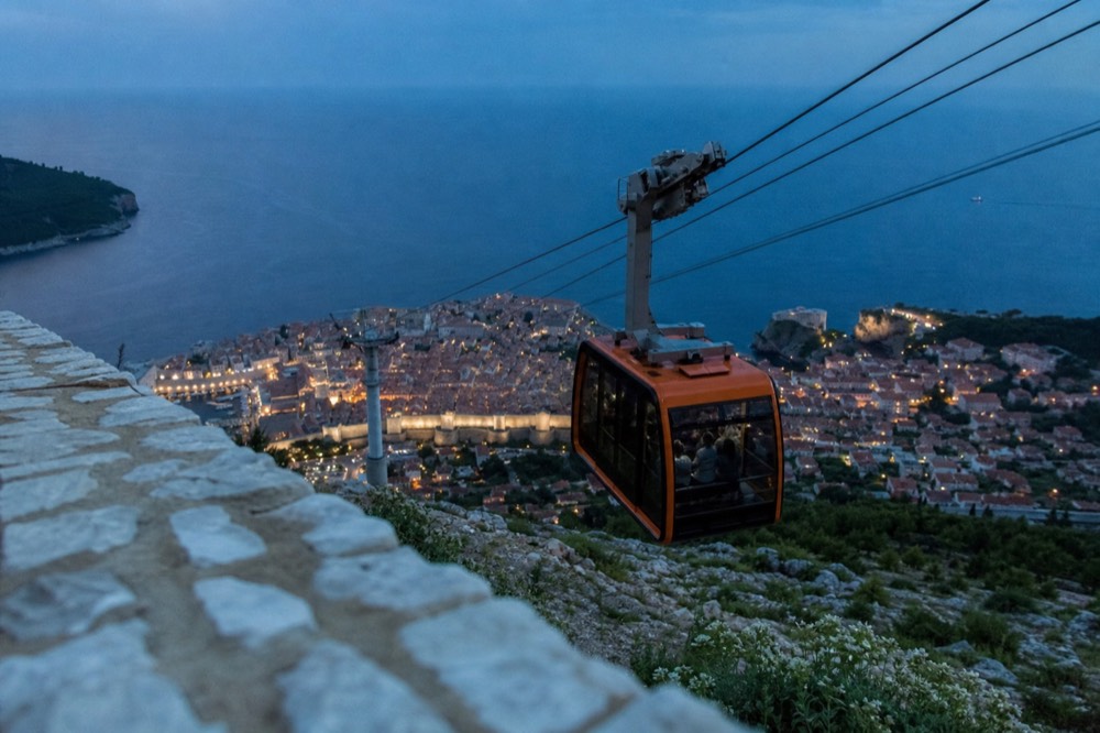 A cable car cabin travelling up a steep hillside above a walled city and the sea