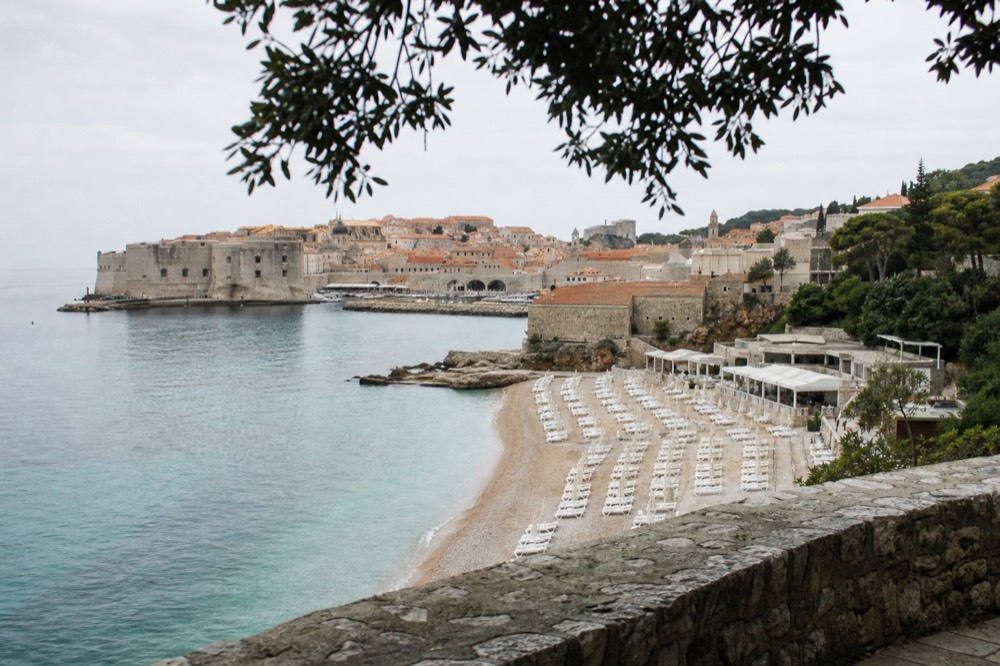 A pebble beach with rows of empty sunbeds and calm blue water, an old town with towers behind it