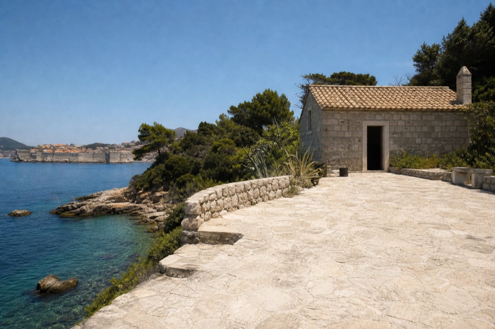 A small stone building above a quiet cove with clear water and a distant walled town