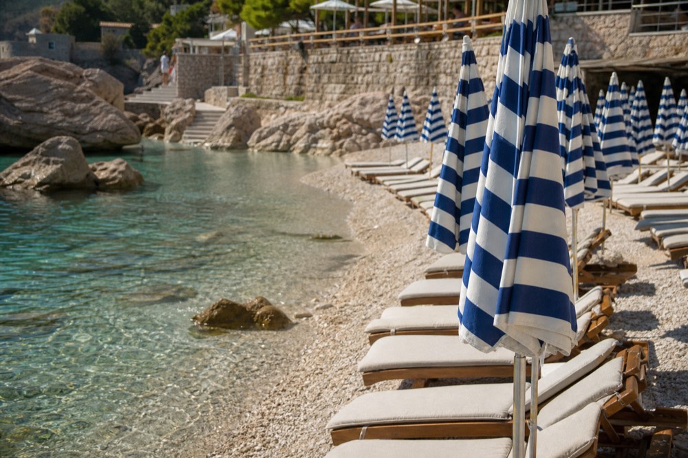 Rows of sunbeds with striped parasols on a pebble shore beside clear shallow water